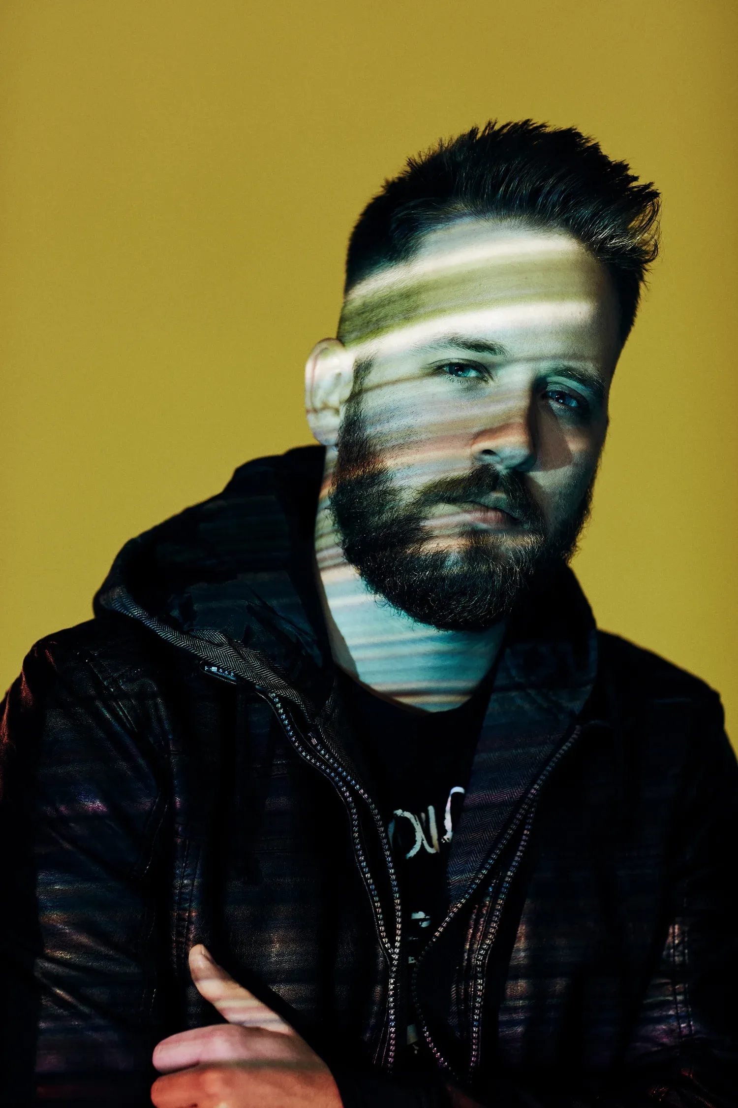 Creative male portrait with intentional motion blur effect across face, styled dark hair and full beard, wearing black leather jacket, photographed against warm golden yellow background, artistic experimental photography retouched by Pratik Naik for photographer Jeremy Cowart