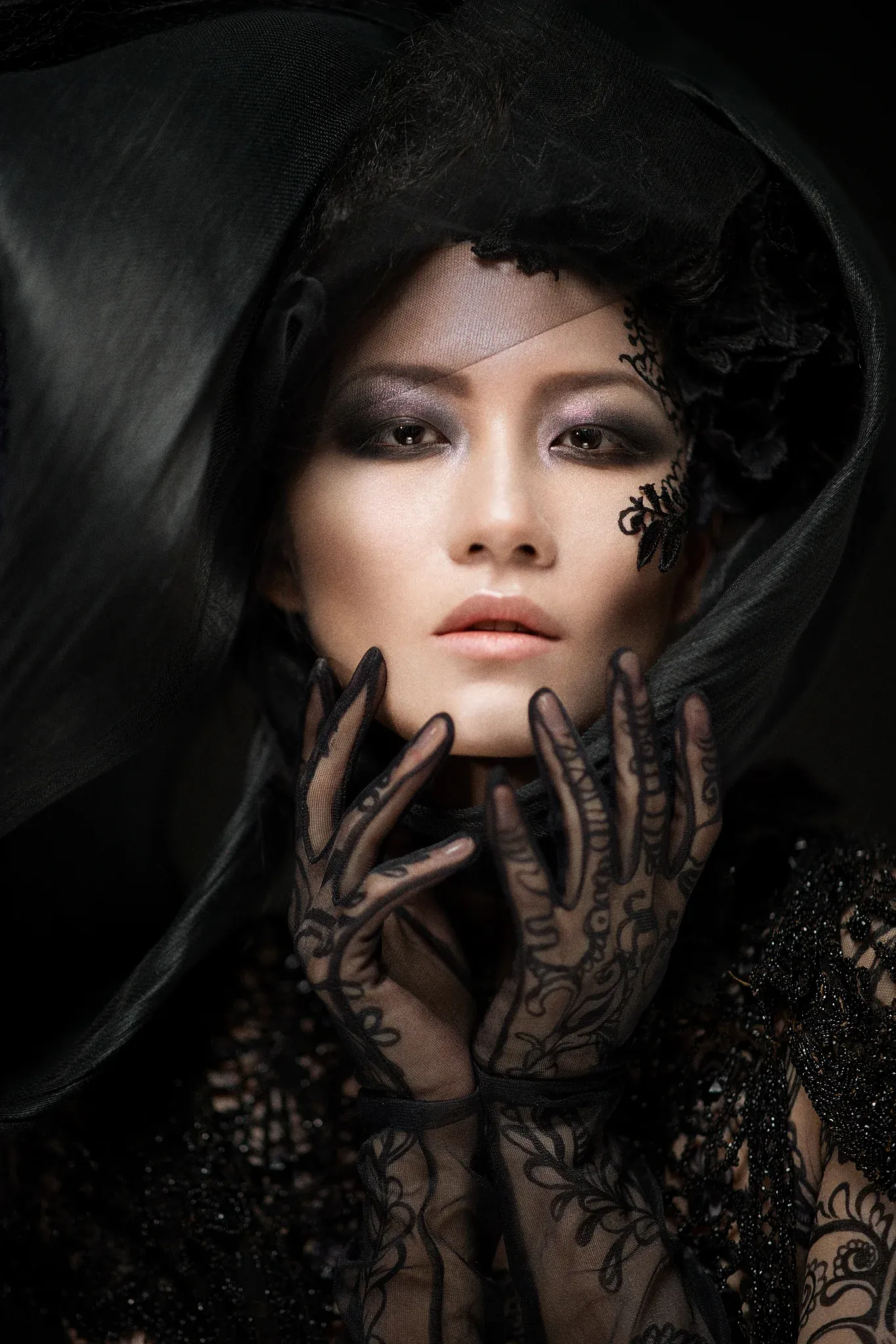 Dramatic beauty portrait of Asian female model with black lace veil and intricate black lace detailing around face and hands, smoky eye makeup, sculpted high-fashion styling on dark background, professional editorial retouching by Pratik Naik for photographer Joe McNally