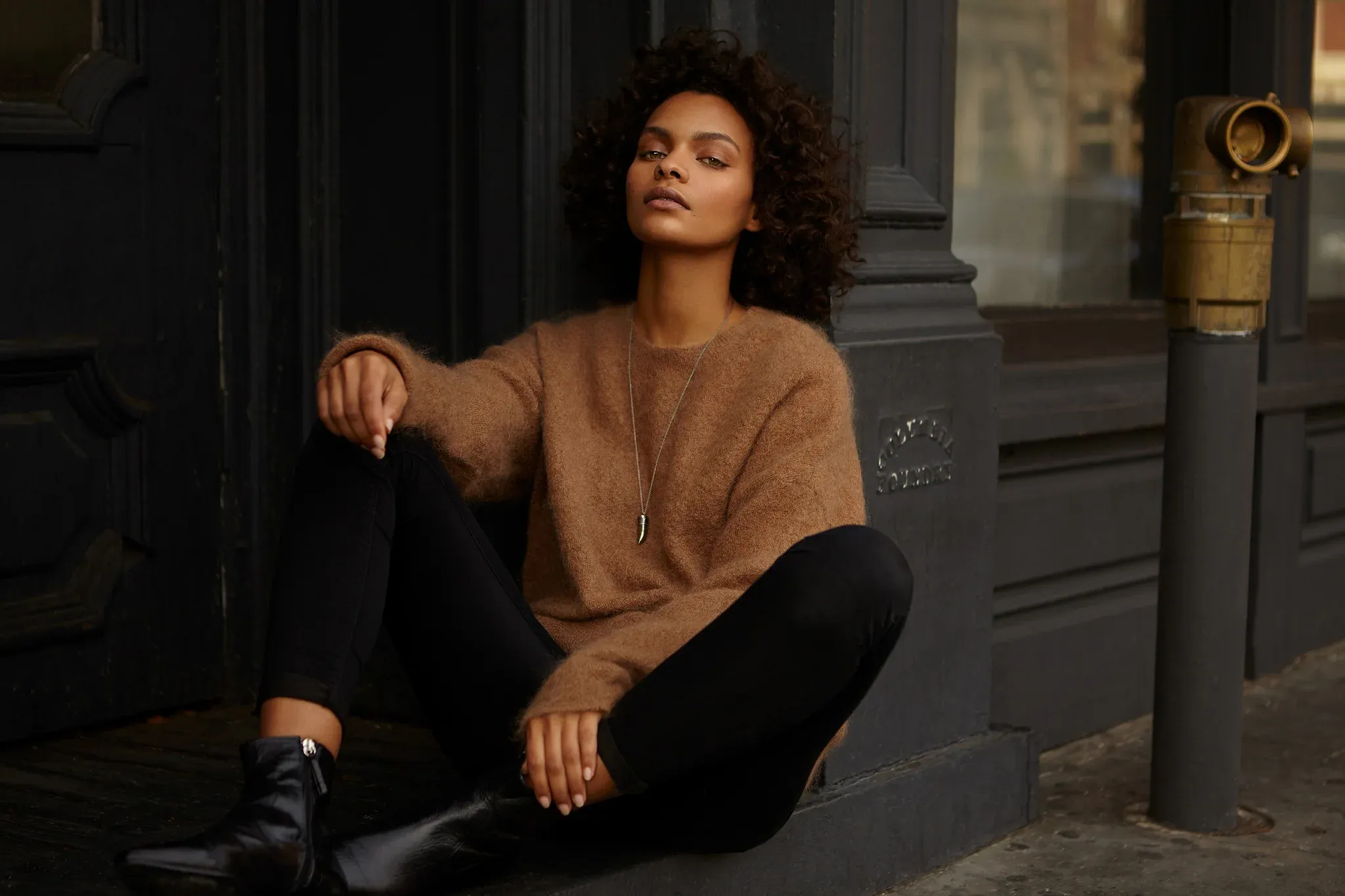 Fashion lifestyle portrait of Black female model with voluminous curly hair wearing tan camel colored sweater and black pants with pendant necklace, relaxed seated pose on urban grey steps with architectural columns, natural golden hour lighting, professional retouching by Pratik Naik for photographer Tricia Turner