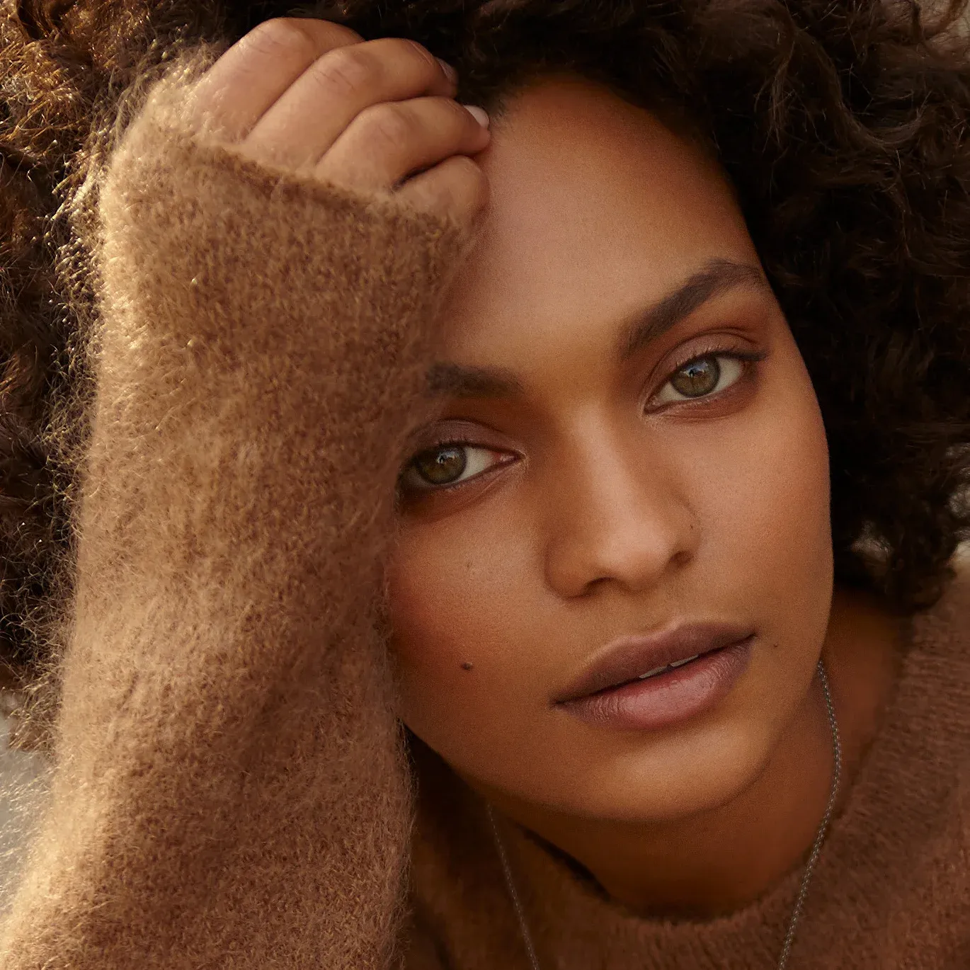 Natural beauty close-up portrait of Black female model with voluminous curly brown afro hair, warm brown eyes, subtle natural makeup, hand touching hair, soft diffused golden lighting, professional skin retouching by Pratik Naik for photographer Tricia Turner