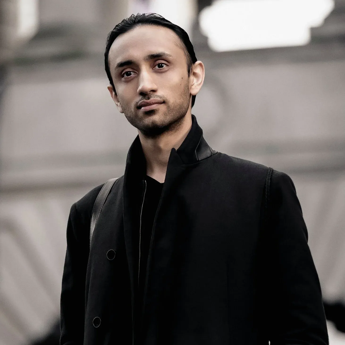 Portrait of male retoucher Pratik Naik with slicked back dark hair, facial hair, wearing all black outfit with black jacket, photographed with soft grey blurred environmental background, professional portrait photography