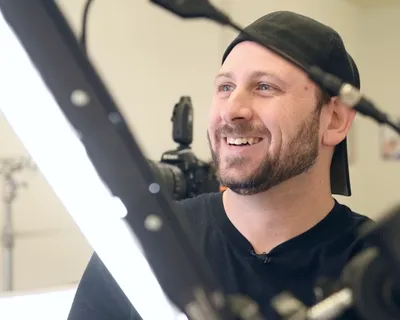 Behind-the-scenes photograph of headshot photographer Richard Waine wearing black baseball cap and black shirt with lavalier microphone attached to collar smiling during headshot session with camera and white studio lighting visible in foreground at Lancaster Pennsylvania studio in 2017