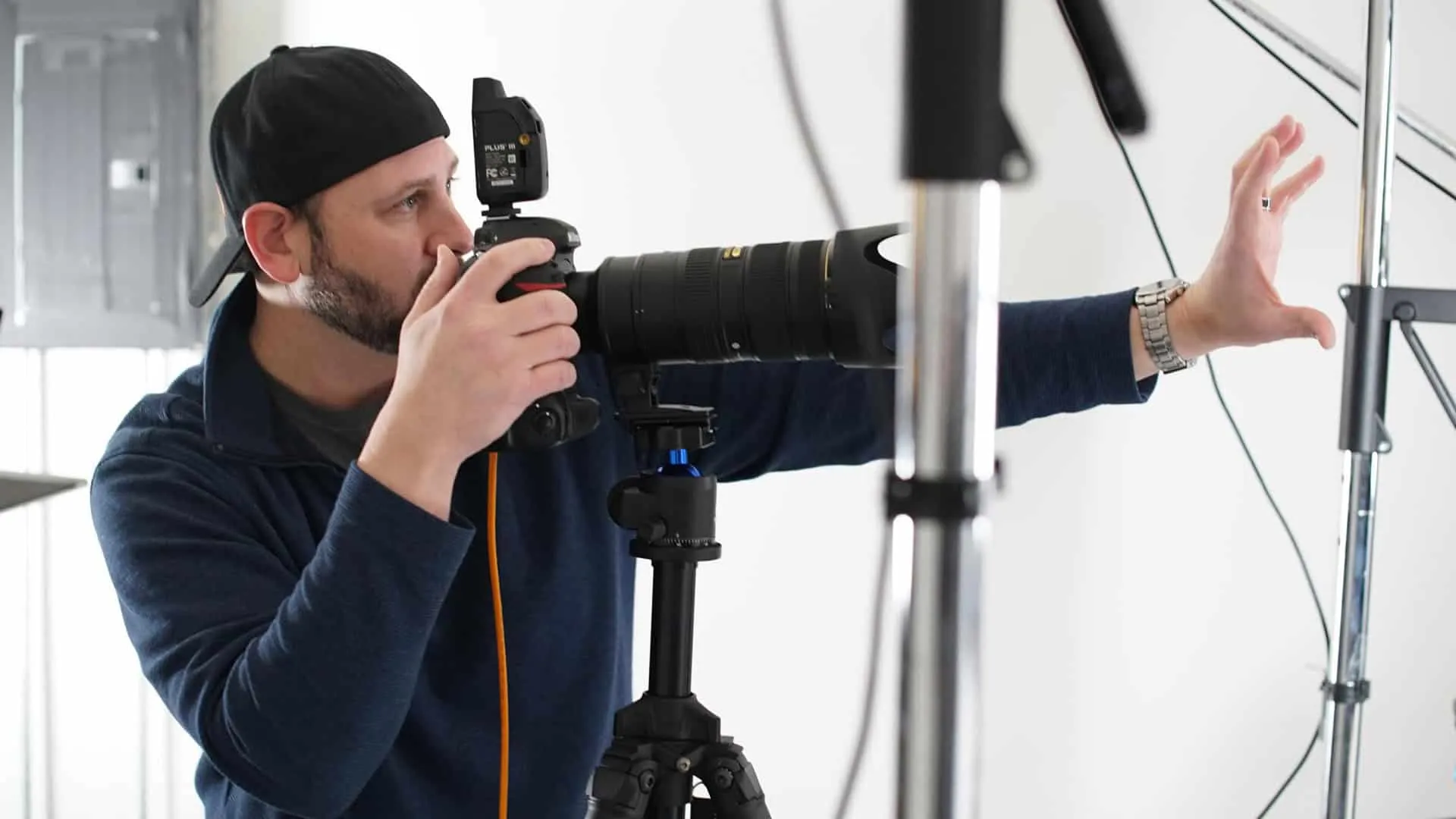Behind-the-scenes photograph of photographer Richard Waine wearing black baseball cap turned backwards and navy blue long-sleeve shirt with trimmed beard looking through professional DSLR camera viewfinder mounted on sturdy black tripod with large telephoto lens attached and wireless flash trigger visible while directing headshot session in bright white photography studio with professional lighting equipment including light stands positioned throughout space at Richard Waine Photography Lancaster Pennsylvania