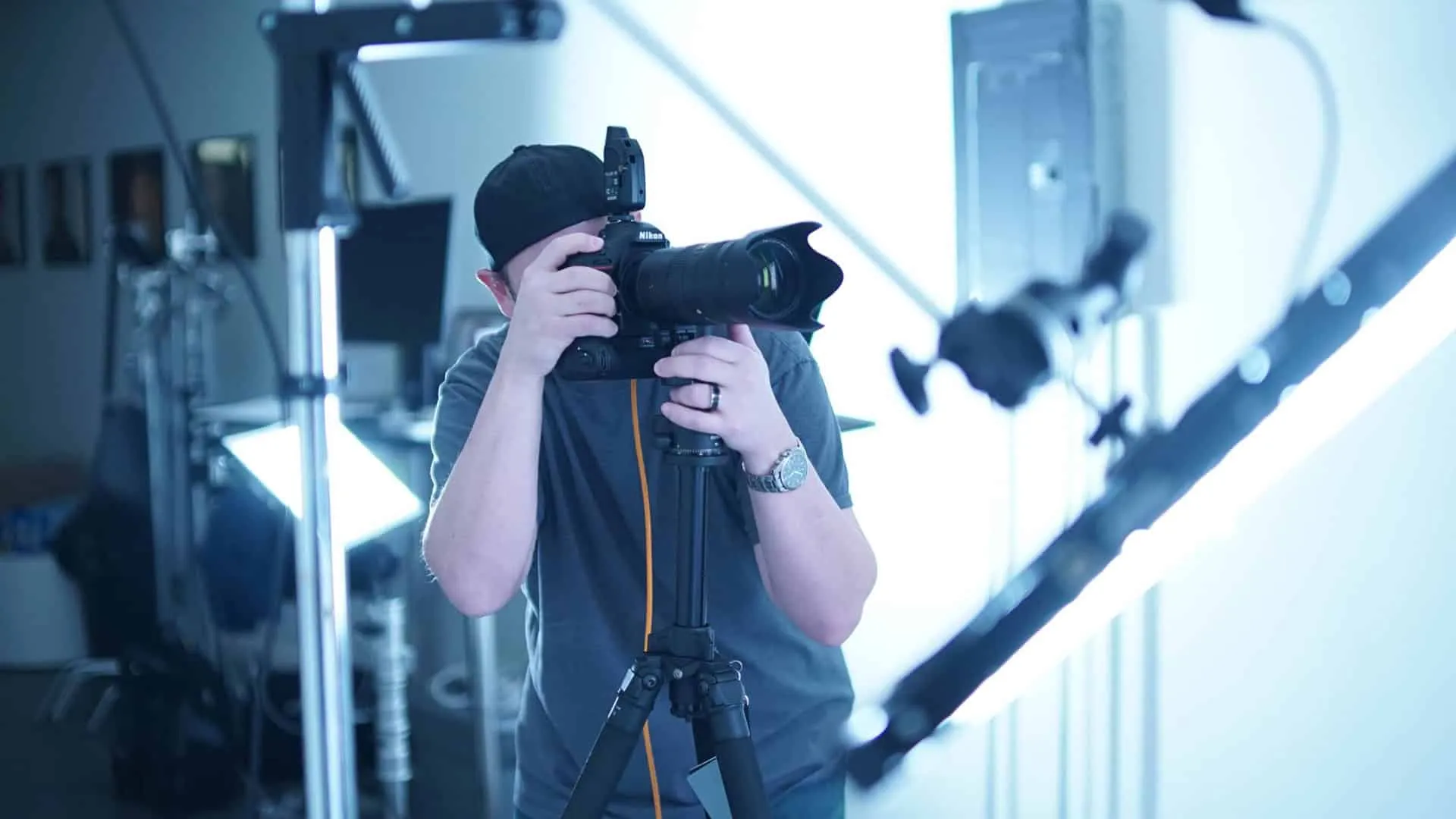 Behind-the-scenes photograph of photographer Richard Waine wearing black baseball cap turned backwards and gray shirt holding professional Nikon DSLR camera with large lens and wireless flash trigger mounted on black tripod while composing shot with both hands on camera wearing wristwatch on left wrist in bright white photography studio with professional lighting equipment and light stands visible in background with cool blue creative color grading tone applied to image at Richard Waine Photography Lancaster Pennsylvania