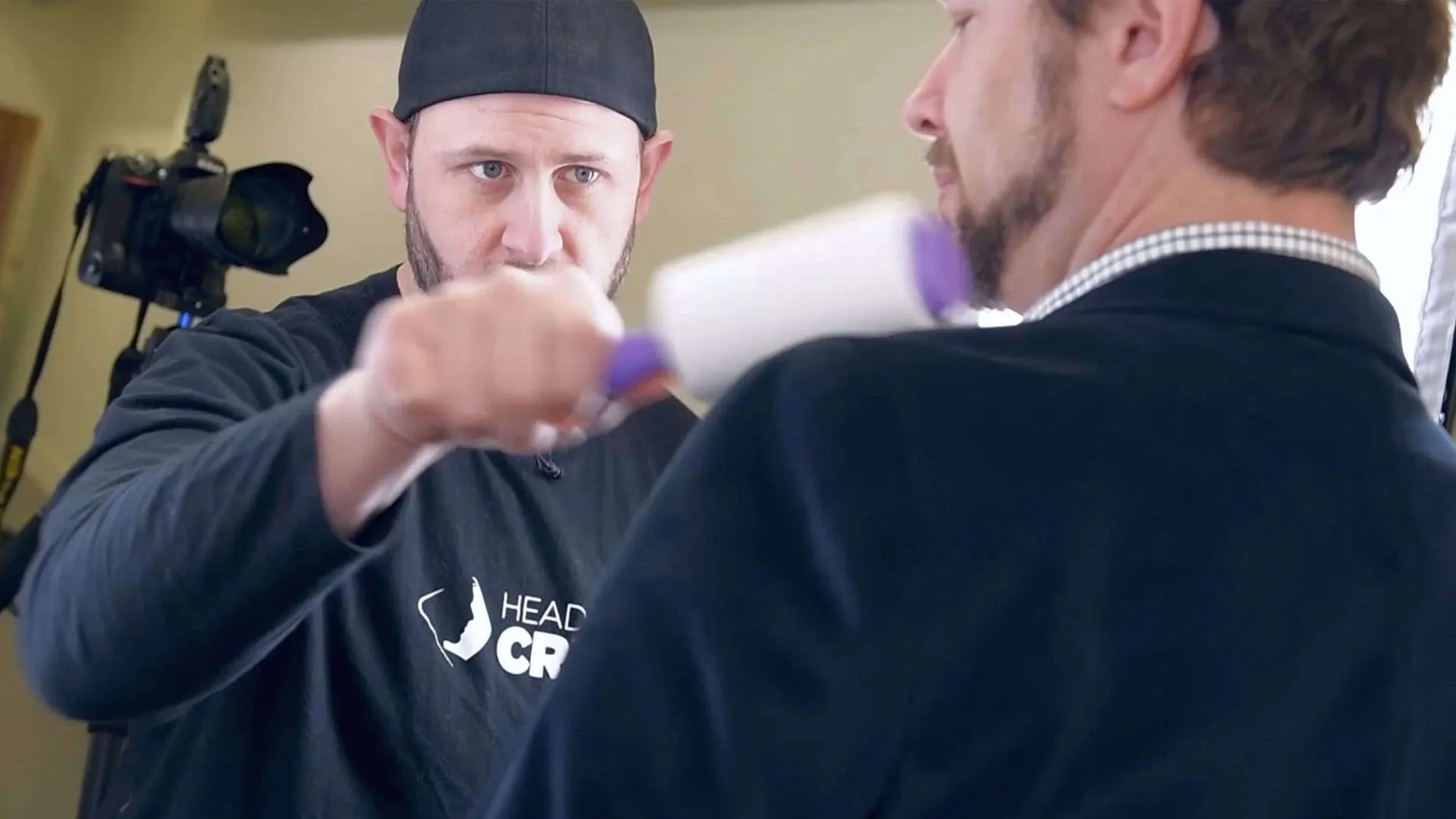 Behind-the-scenes photograph of photographer Richard Waine wearing black baseball cap turned backwards and black long-sleeve shirt using purple lint roller to remove lint from male client's black clothing while client shown from behind wearing black vest over checkered shirt with professional photography equipment and lighting visible in background demonstrating professional presentation and client preparation process at Richard Waine Photography Lancaster Pennsylvania