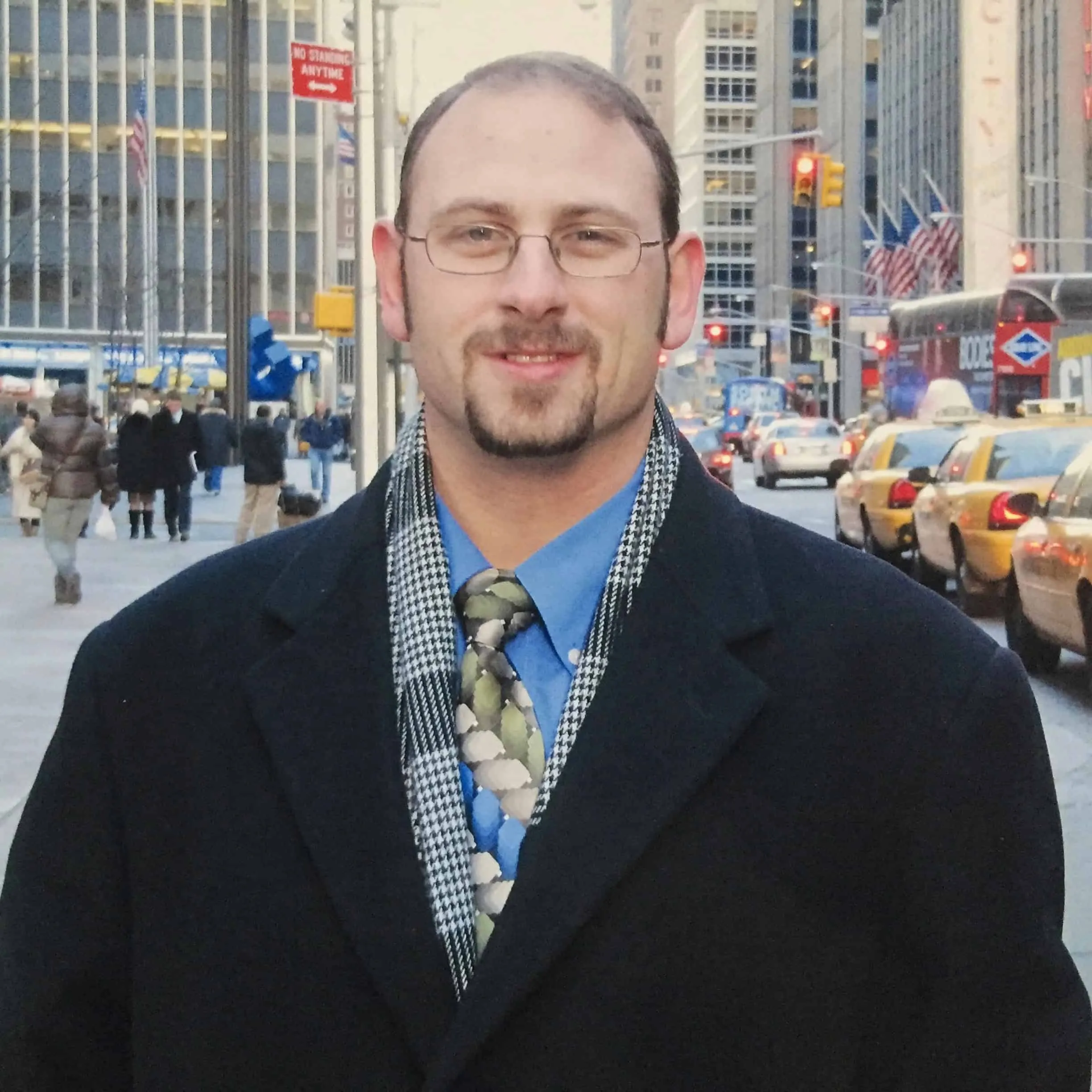 Vintage professional photograph of young Richard Waine wearing wire-rimmed eyeglasses black wool overcoat blue dress shirt patterned necktie and black and white houndstooth scarf standing on busy New York City street with pedestrians yellow taxi cabs and tall urban office buildings visible in background from Wall Street career days demonstrating professional business appearance and style