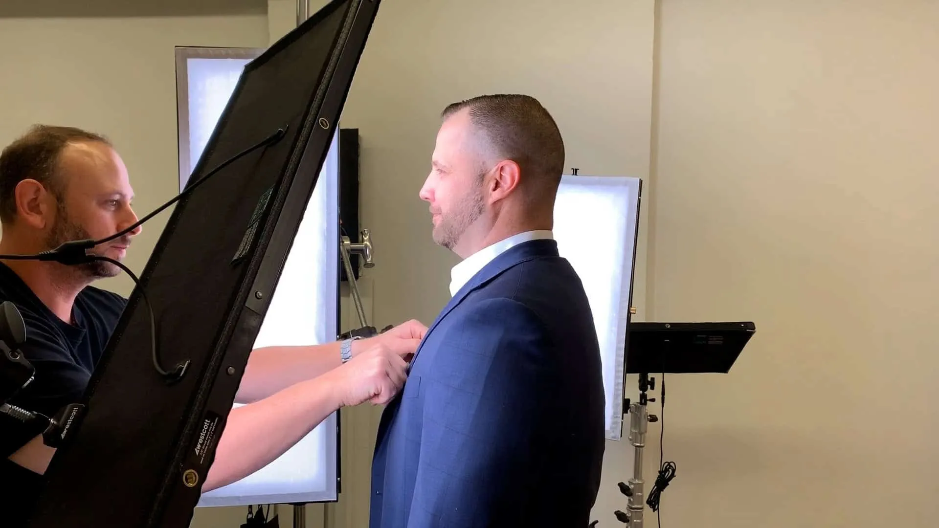 Behind the scenes headshot photography session showing photographer Richard Waine in black shirt with brown hair and beard adjusting navy blue suit on male client in profile view, standing in front of white LED panel lights on stands in Lancaster Pennsylvania studio, professional headshot production process