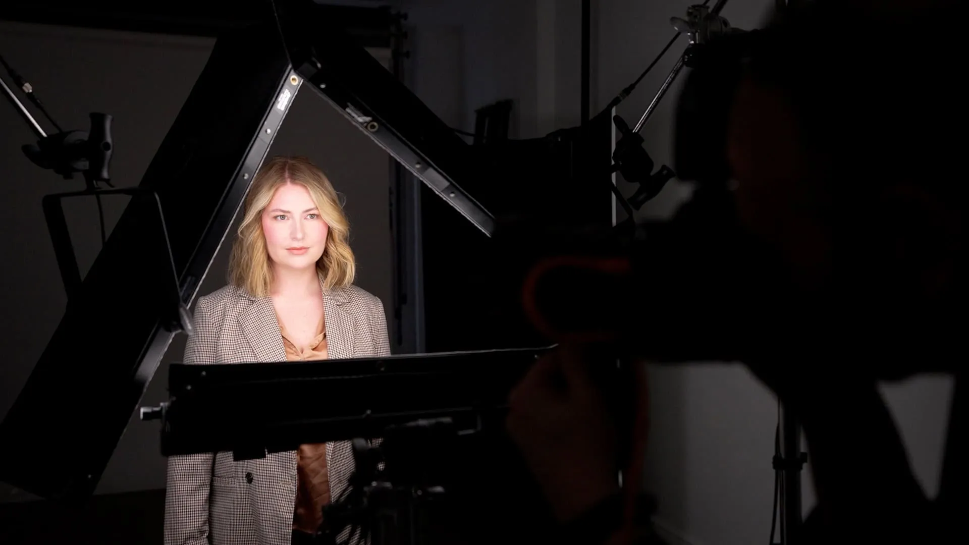 Behind the scenes headshot photography session showing female subject with shoulder-length wavy blonde hair wearing tan houndstooth pattern blazer standing in front of black continuous light panels on stands in Lancaster, Pennsylvania headshot photography studio