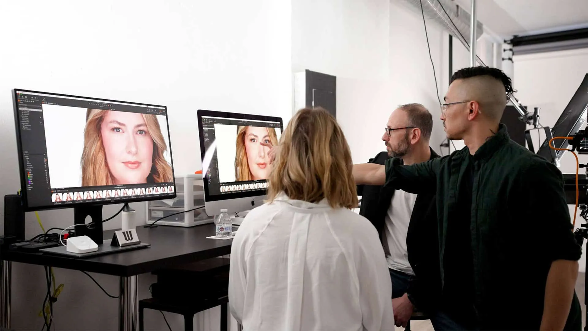 Behind the scenes headshot image review session showing female client in white top seated at desk facing two computer monitors displaying headshot images, photographer Richard Waine with brown hair and beard in eyeglasses pointing at screen, makeup artist with short black styled hair wearing dark green shirt standing together reviewing images in Lancaster, Pennsylvania headshot photography studio