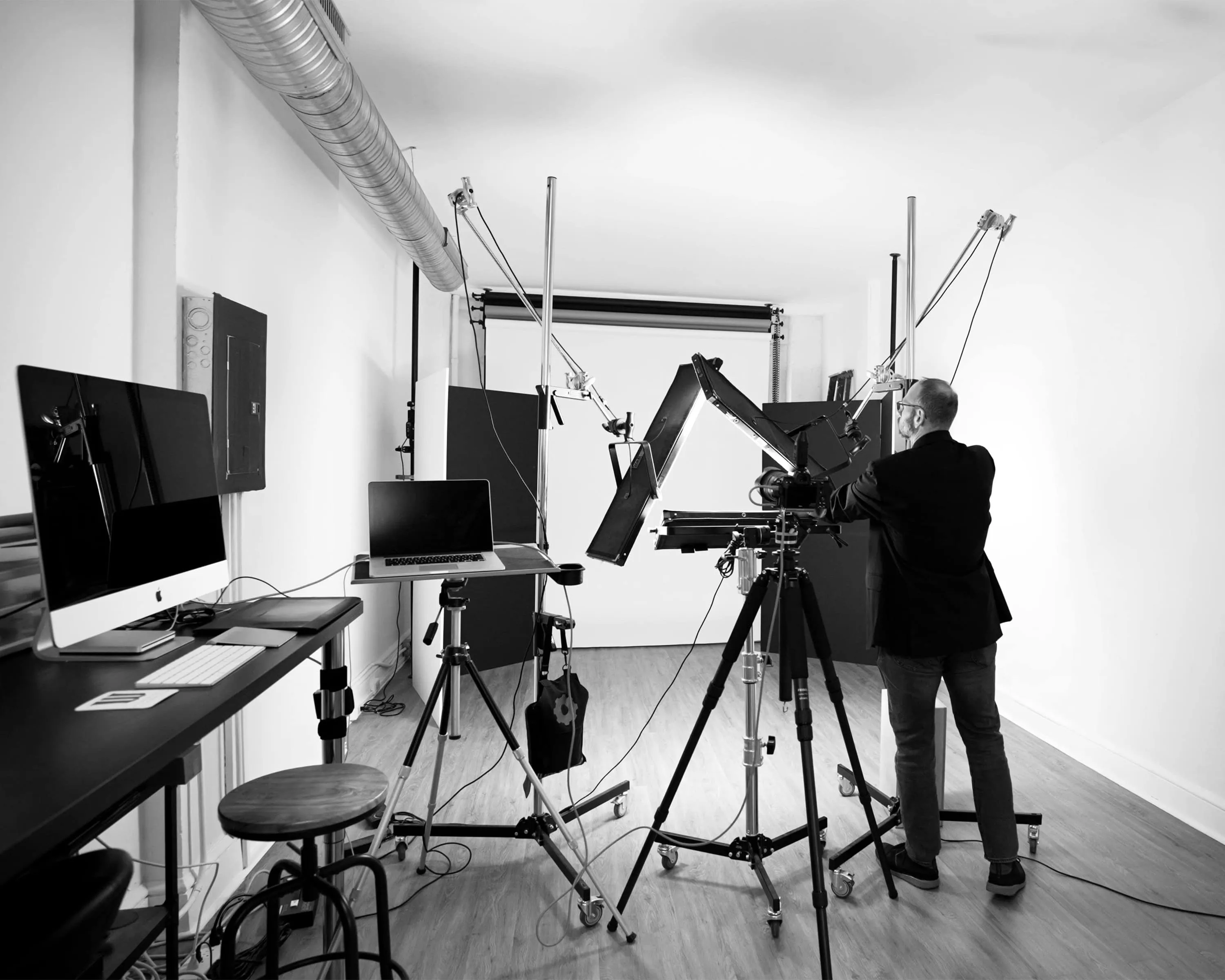 Black and white behind-the-scenes photograph of photographer in business attire arranging professional studio lighting equipment stands and white seamless backdrop with exposed industrial ceiling details at Richard Waine Photography Lancaster Pennsylvania headshot studio