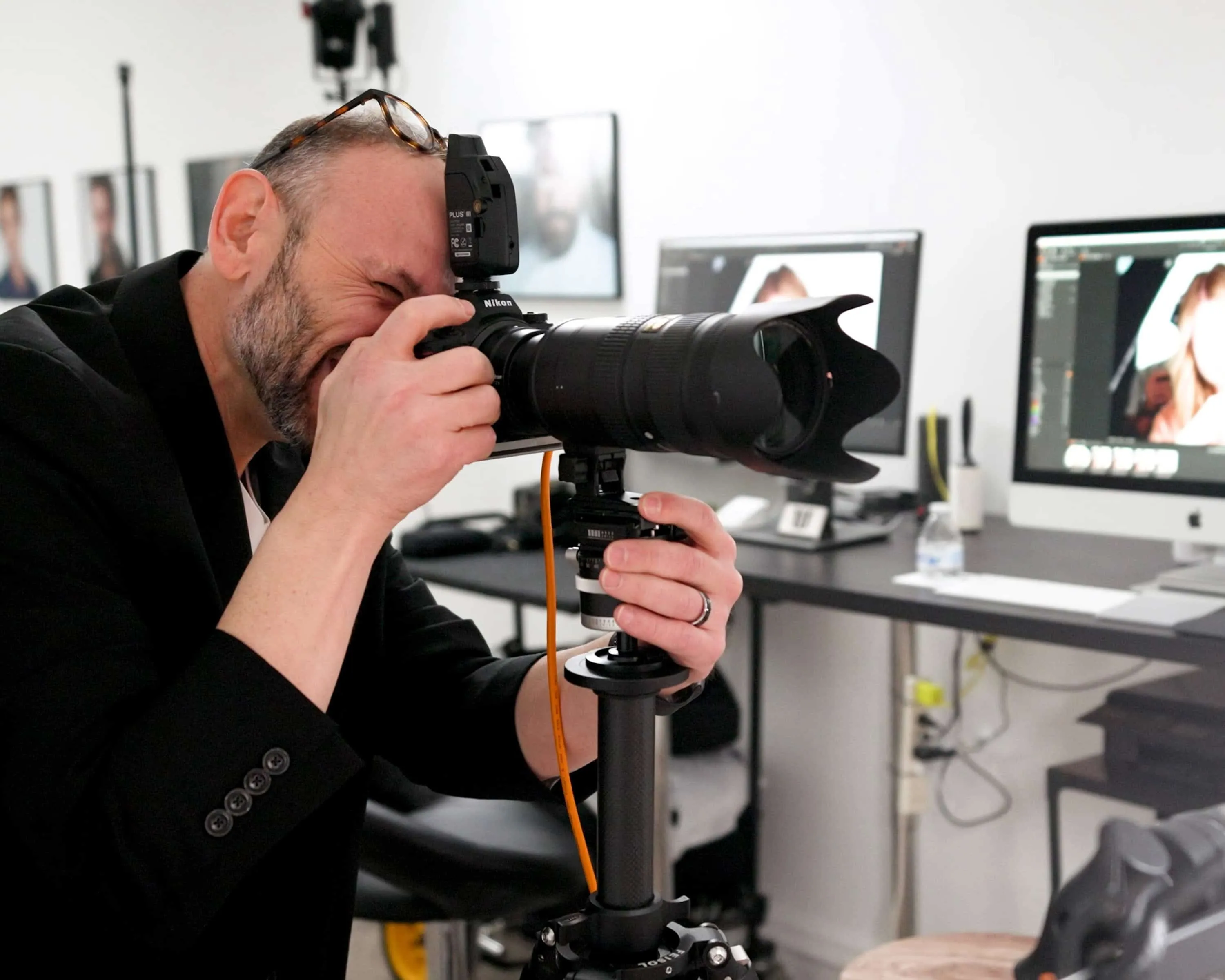 Photographer Richard Waine with trimmed beard and eyeglasses resting on top of head carefully composing shot through professional camera viewfinder with computer monitors displaying headshot images visible in background at Lancaster Pennsylvania photography studio