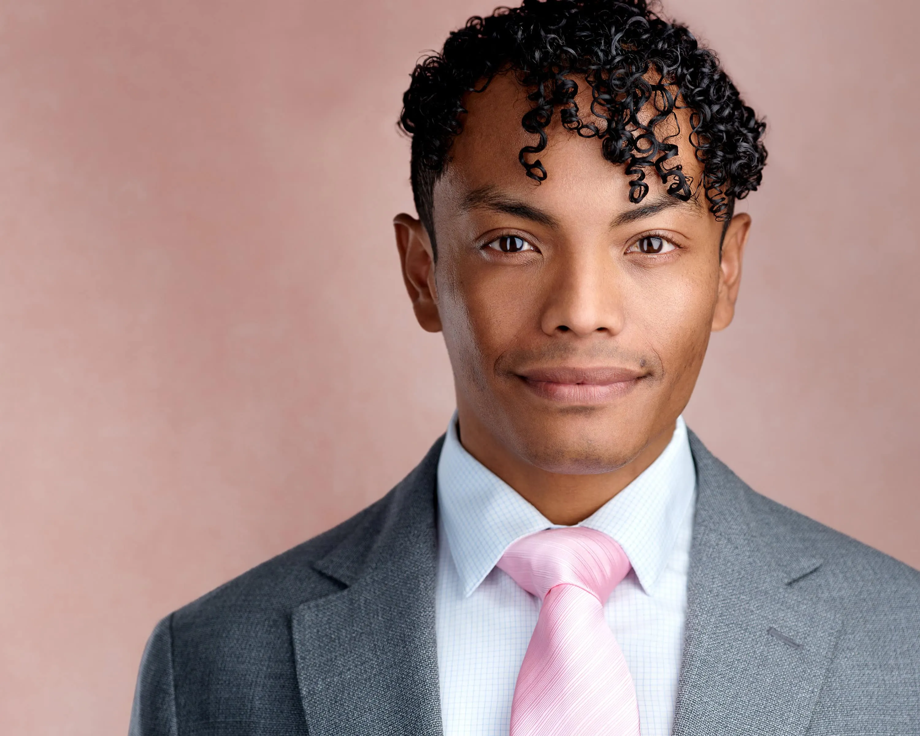 Professional headshot of male with short black curly hair, wearing charcoal grey suit jacket over light blue dress shirt with pink diagonal striped tie, subtle smile, photographed on pink canvas background in Lancaster, Pennsylvania by headshot photographer Richard Waine