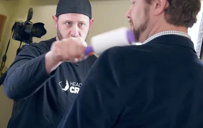 Behind-the-scenes photograph of headshot photographer Richard Waine wearing black baseball cap using lint roller to prepare male client's clothing during professional headshot session with photography equipment visible in background at Lancaster Pennsylvania studio