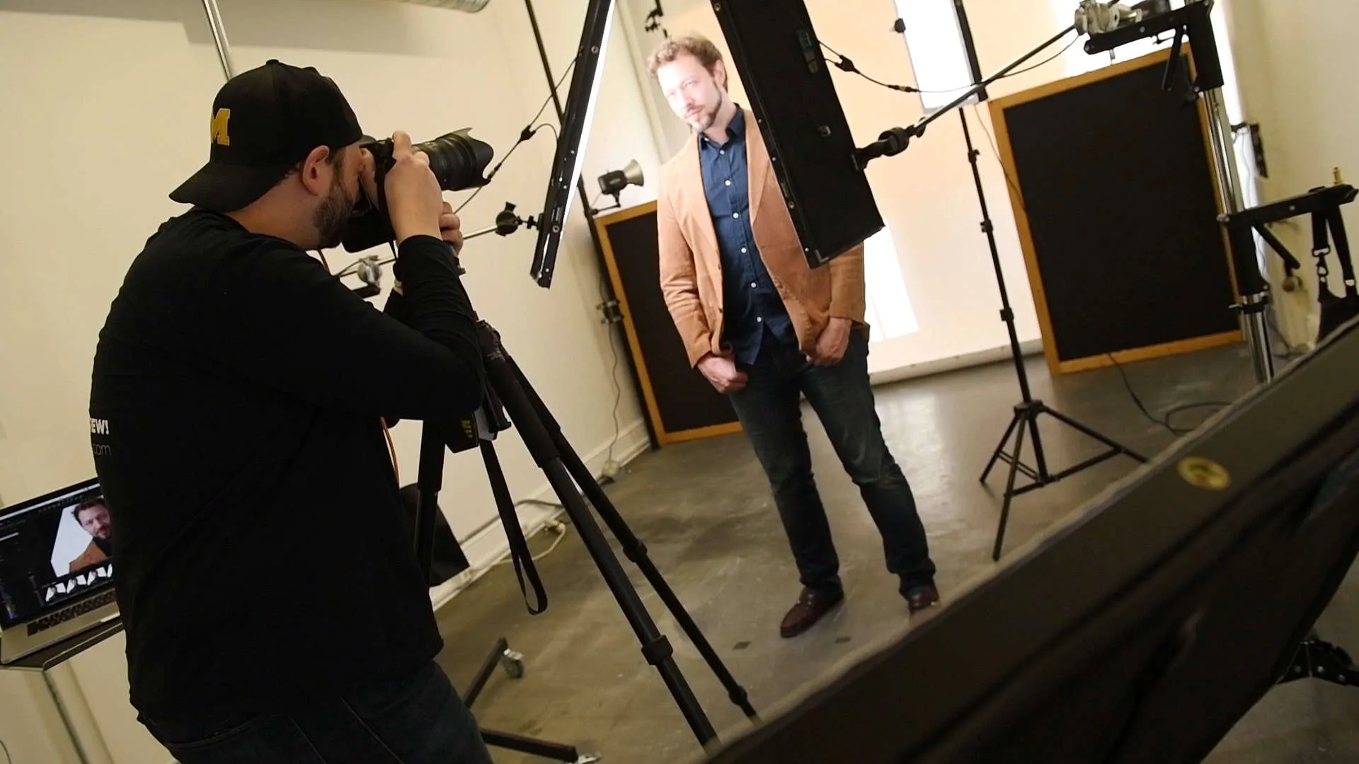 Behind the scenes studio photography session showing photographer Richard Waine in black shirt and black baseball cap looking through viewfinder of DSLR camera mounted on tripod, male subject in tan blazer over navy shirt and blue jeans standing on seamless white backdrop, two black studio strobes on light stands aimed at white background, LED panels illuminating subject, laptop on table in foreground, Lancaster Pennsylvania headshot photography studio setup