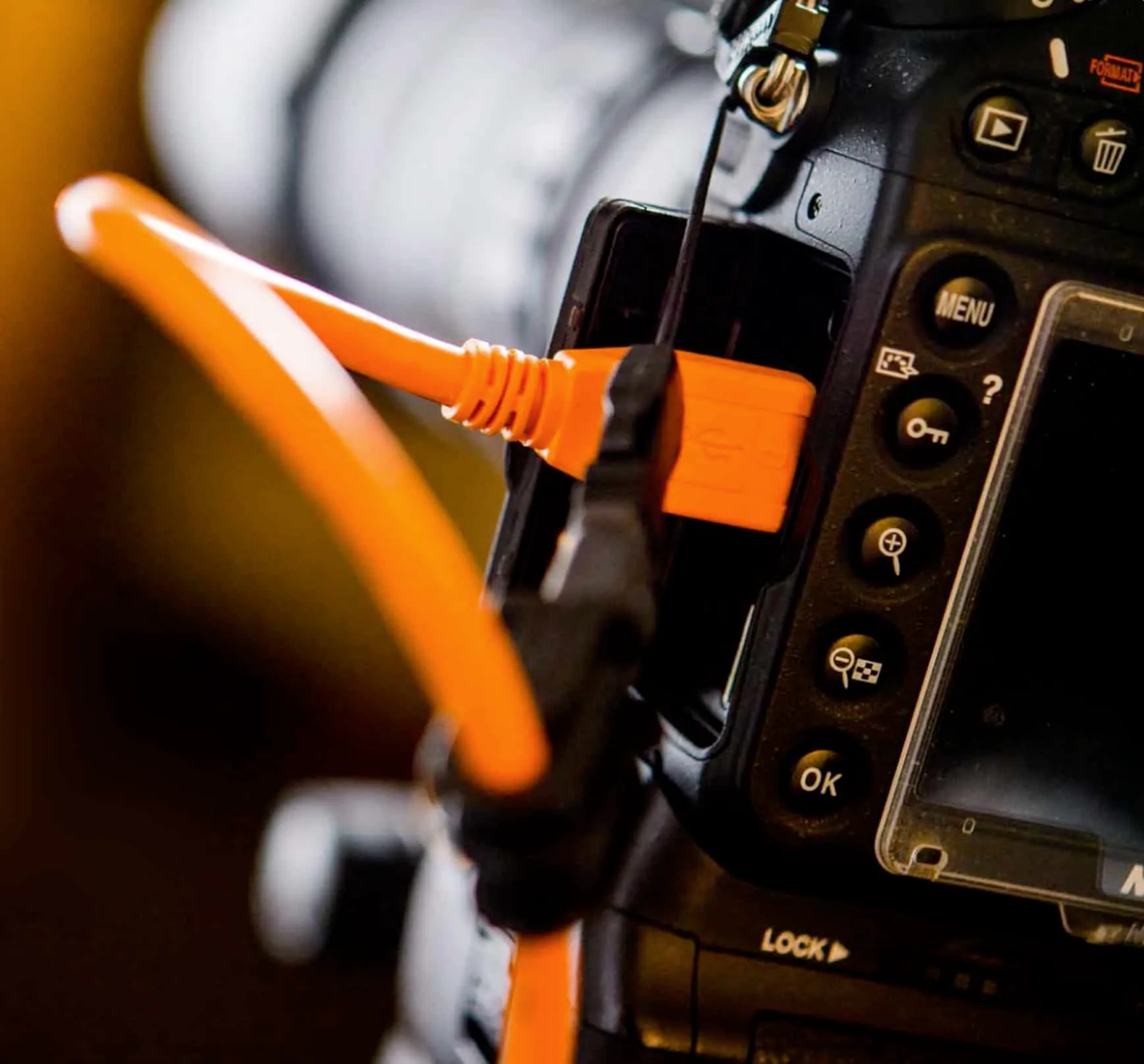 Close-up macro photograph of bright orange TetherTools USB tether cable plugged into side port of black Nikon DSLR camera body, camera control buttons including menu ok lock visible, cable strain relief connector, shallow depth of field with blurred background, professional tethering equipment for studio photography workflow